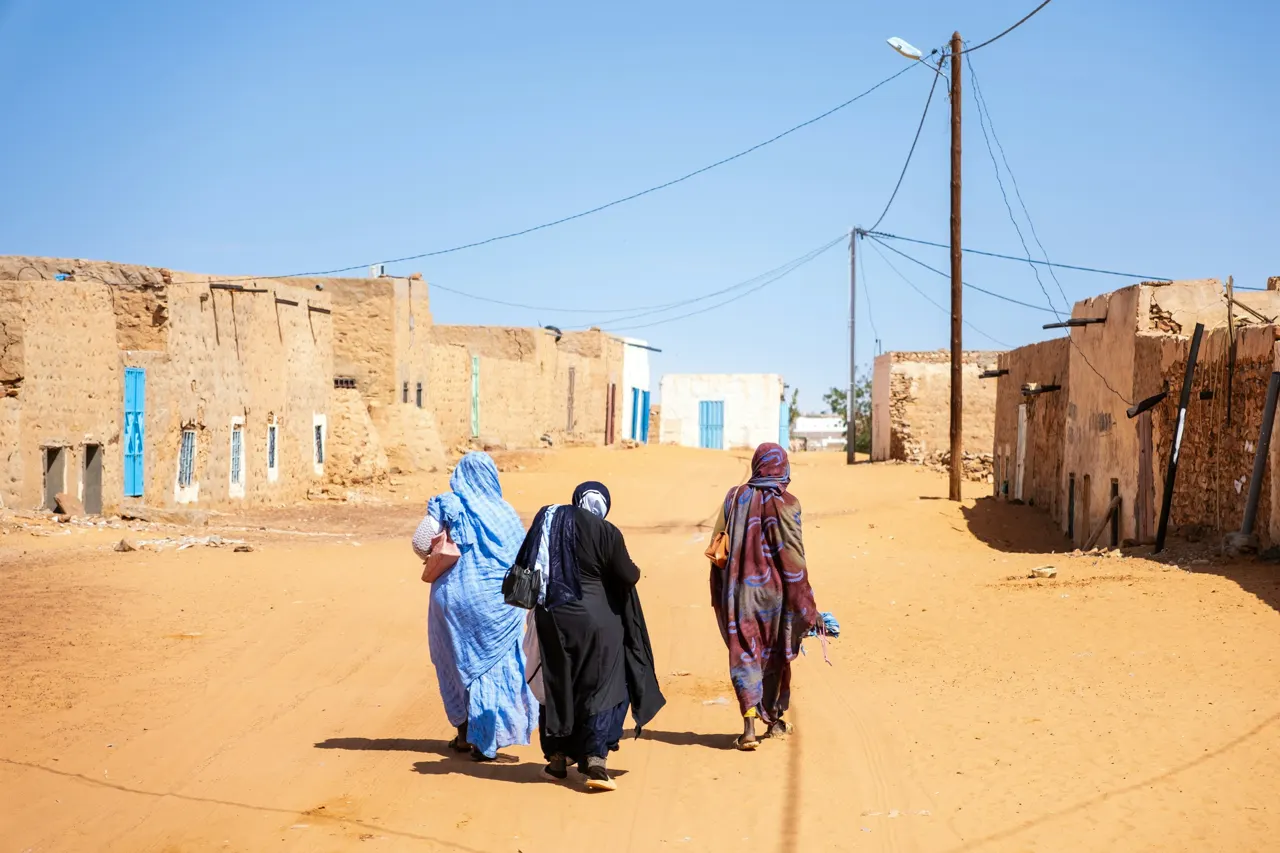 Candid street photography capturing three women in traditional melhafas walking down a sun-drenched, sandy street lined with adobe houses in Mauritania