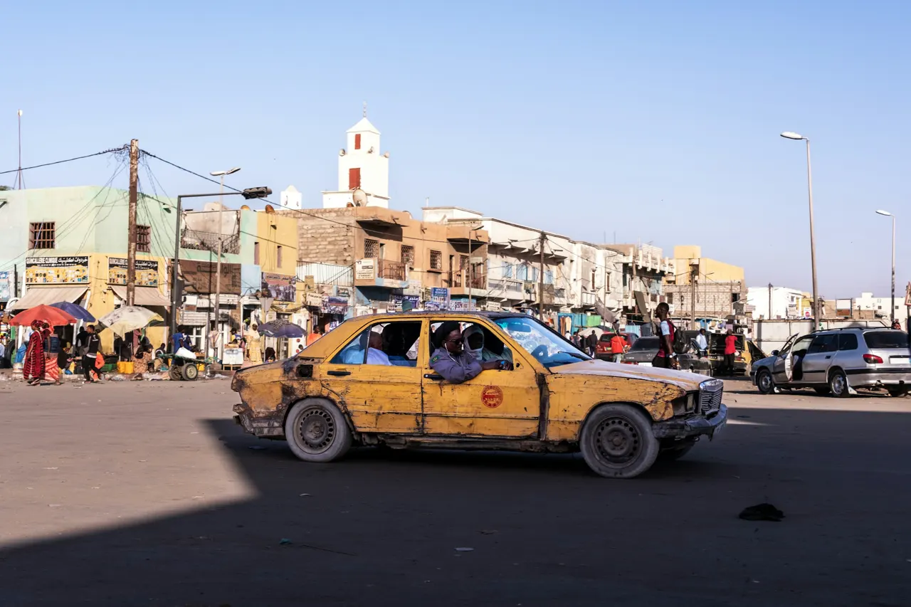 Authentic street photography in Nouakchott, Mauritania, featuring a weathered yellow vintage taxi carrying passengers across a dusty city square under a bright desert sky