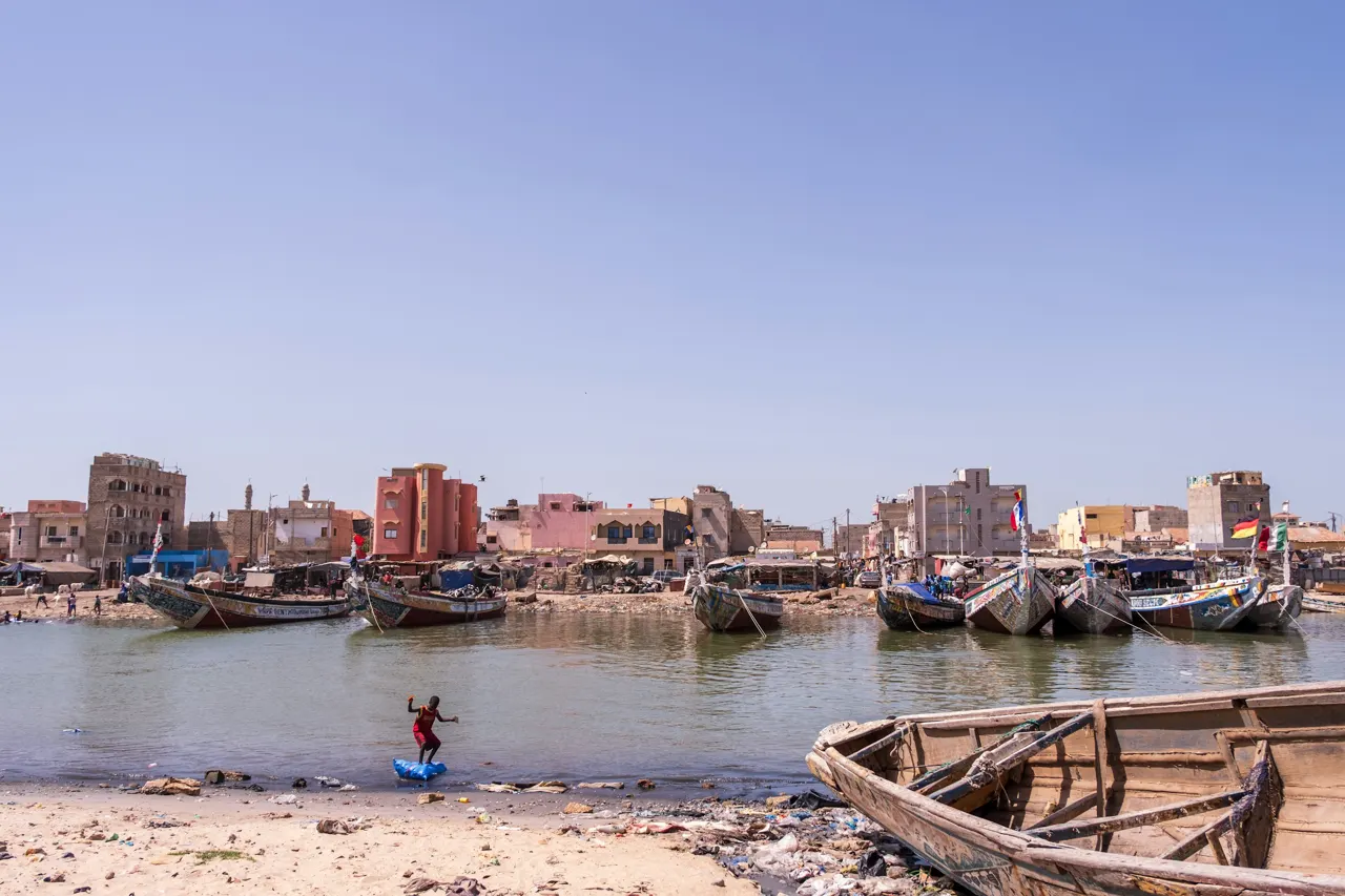 This wide-angle photograph captures a vibrant waterfront scene in Saint-Louis, Senegal, under a clear, expansive sky