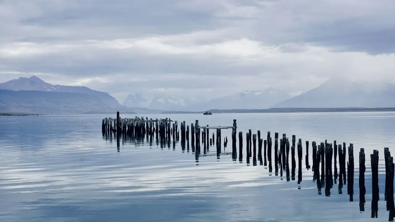 A weathered wooden pier extending into a calm lake beneath distant snow-capped mountains