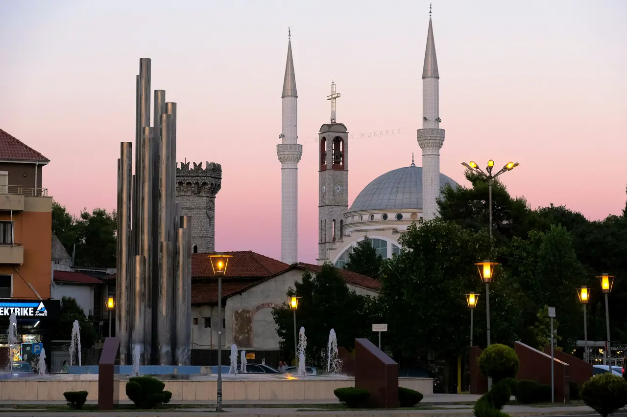 A striking urban scene at dusk, featuring a mosque with two towering minarets, a historic stone tower, and a modern metallic monument in the foreground